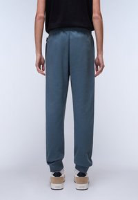 Lightweight, tapered joggers in slate blue. Features an elastic waistband and side pockets. Paired with beige and white sneakers.