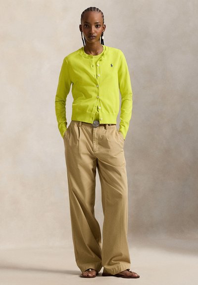 Λεπτή ζακέτα σε πράσινο χρώμα λάιμ με κουμπιά μπροστά, συνδυασμένη με μπεζ παντελόνες φαρδιάς γραμμής. Διακριτική ασημένια αγκράφα ζώνης και casual σανδάλια.