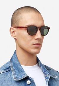 Kerek napszemüveg teknőspáncél mintás kerettel és sötét lencsékkel, egy farmerkabátot és fehér inget viselő személyen világos háttér előtt.