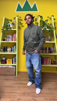 Chaqueta verde con cremallera y logo, vaqueros azules desgastados y zapatillas blancas. Fondo con pared amarilla, estantes verdes, libros y plantas.