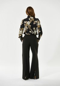 Schwarze Bluse mit floralem Muster und Rüsche am Kragen, kombiniert mit hochgeschnittenen schwarzen Hosen mit seitlichen Schlitzen. Das Outfit hat eine glatte Textur.