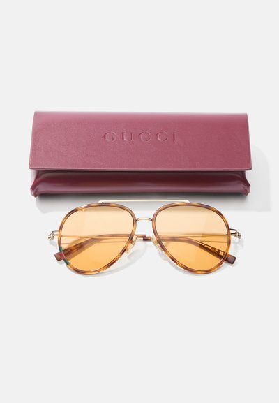 Gafas de sol con tinte marrón y marco de metal, con una forma ovalada distintiva. Viene con un estuche de cuero burdeos grabado con "Gucci."