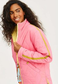 Prošívaná růžová bunda se žlutou podšívkou, s zelenými akcenty a dekorativním táhlem zipu. Design zahrnuje vysoký límec a postranní kapsy.
