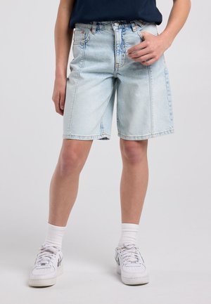 Personne portant un short en jean bleu clair jusqu'aux genoux, des baskets blanches avec des chaussettes et un haut foncé sur un fond uni.