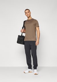 Chemise marron à manches courtes, pantalon noir et baskets blanches. Le mannequin tient un sac noir avec des accents en maille. Fond uni, couleurs neutres.