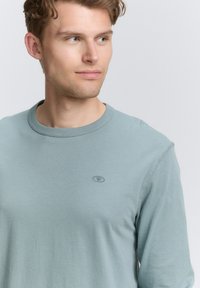 Himmelblaues Langarm-T-Shirt aus weicher Baumwolle. Runder Halsausschnitt mit genähten Kanten, mit einem kleinen Logo auf der linken Brust.