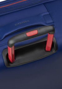 Valigia blu con ruote in primo piano, che mostra un manico retrattile rosso con impugnatura nera, tessuto testurizzato e zip arancione a contrasto.