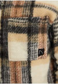 Koszula w kratę z polaru z naszywaną kieszenią, dostępna w kolorach: beżowym, czarnym i pomarańczowym. Teksturowany materiał w wzór szachownicy. Widoczna czarna metka.