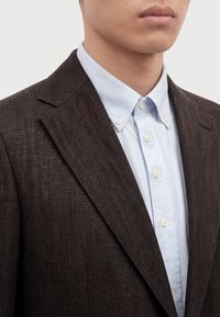Homme portant un blazer marron foncé texturé sur une chemise bleu clair boutonnée avec un col boutonné, montré des épaules aux lèvres.