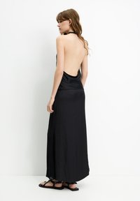 Schwarzes Satin-Maxi-Kleid mit offenem Rücken, lockerer Passform, seitlichen Schlitzen und dezenten Spitzenakzenten am Saum. Flache schwarze Sandalen dazu getragen.