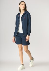 Completo sportivo navy composto da una felpa con cappuccio con zip, pantaloni coordinati e sneakers bianche, con un piccolo logo rosso sul petto e sui pantaloni.