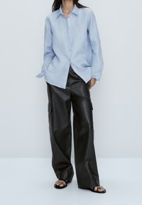 Camicia button-up azzurro chiaro realizzata in lino, abbinata a pantaloni cargo neri in pelle con tasche laterali e vestibilità a gamba larga. Sandali completano il look.