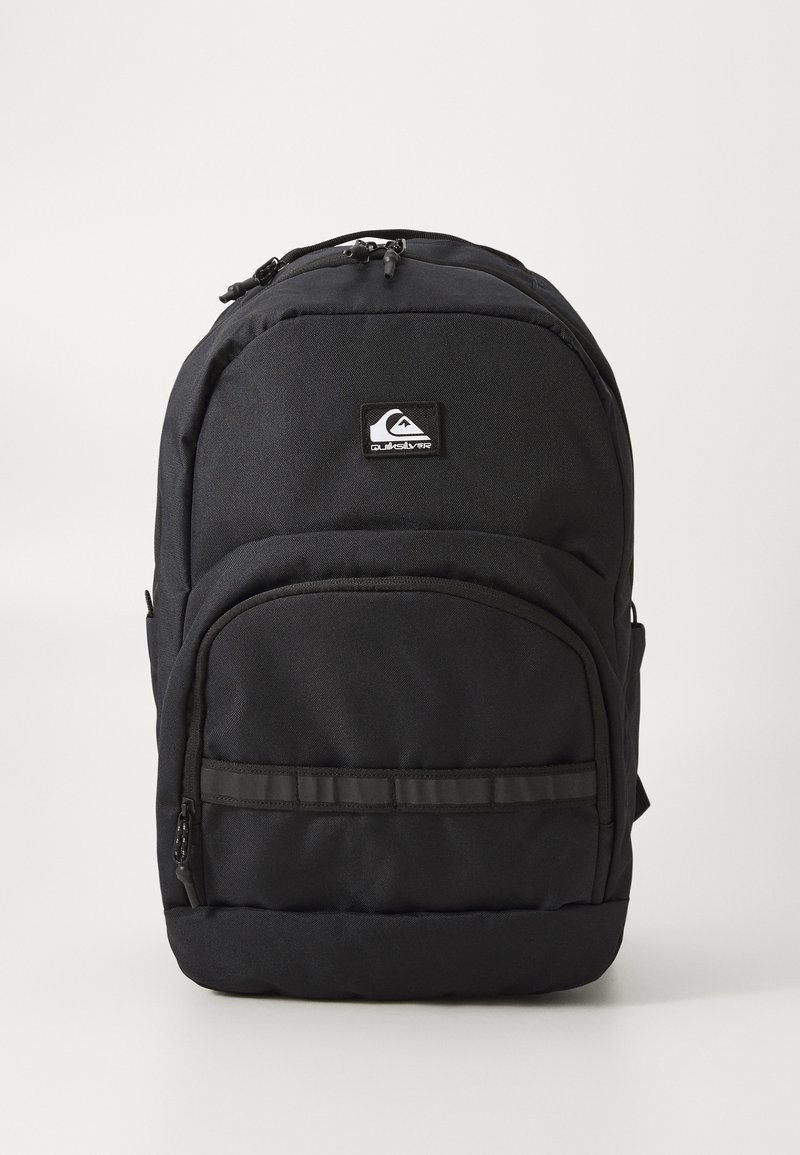 Mochila negra de Quiksilver con bolsillo frontal con cremallera, compartimento principal acolchado y parche con el logo sobre fondo blanco liso.