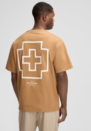 Bruine katoenen t-shirt met een grote witte Zwitserse kruis graphic op de achterkant, met daaronder de tekst "From SWITZERLAND with Love".