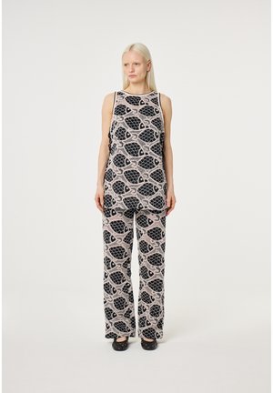 Vrouw met lang blond haar, gekleed in een bijpassende mouwloze top en broek met een zwart vispatroon op een lichte achtergrond, staand op blote voeten.