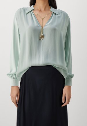 Kvinde iført lys grøn langærmet bluse med åben krave, sort nederdel og et guldhalskæde med hængende bogstavscharme.