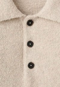 Beżowa wełniana koszula o fakturowanej powierzchni, z ostrym kołnierzem i trzema czarnymi guzikami z przodu.