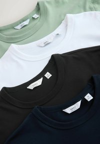 Quatre t-shirts à col rond en vert sauge, blanc, noir et bleu marine, soigneusement superposés, montrant les étiquettes de taille et de marque.