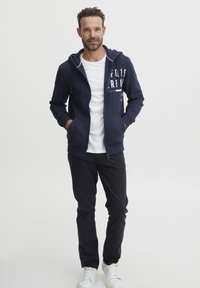 Sudadera con cremallera en color navy con acentos de logo blanco, combinada con una camiseta blanca y pantalones oscuros. El modelo lleva zapatillas blancas y tiene las manos en los bolsillos.