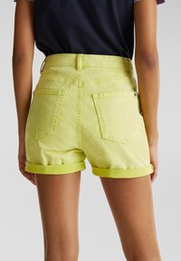 Personne portant un short en denim jaune taille haute avec revers, et une chemise noire rentrée, vue de dos sur un fond uni.