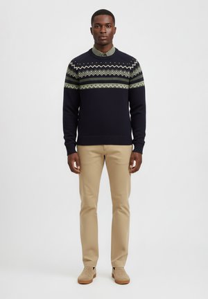 Jersey de color marino con patrón geométrico verde en la parte superior, combinado con pantalones beige claros y calzado beige, de pie sobre un fondo sencillo.