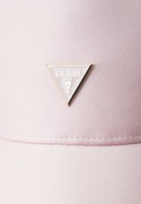 Różowa czapka z teksturowanej tkaniny, z trójkątnym złotym logo i napisem "GUESS USA" oraz wzorem znaku zapytania.