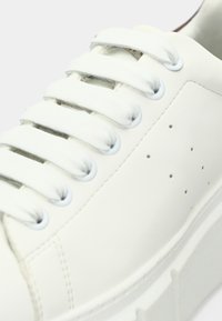 Λευκό δερμάτινο αθλητικό παπούτσι με επίπεδα λευκά κορδόνια, ελάχιστες τρύπες στο πλάι και παχύ λευκό πέλμα. Ομοιόμορφη υφή σε όλο το παπούτσι.