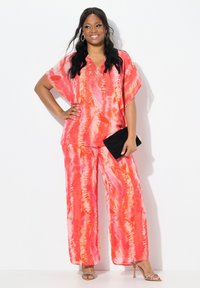 Dvoudílný outfit s volným střihem, tie-dye vzorem v odstínech růžové a oranžové, doplněný černou psaníčkem a sandály na podpatku.