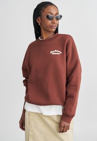 Sweatshirt castanho em algodão com o logótipo branco "New Balance Athletics", gola redonda com nervuras e corte solto, sobreposto a uma camisa branca e uma saia creme.