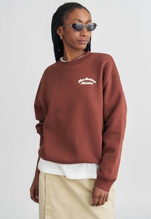 Felpa in cotone marrone con logo bianco "New Balance Athletics", collo a girocollo a costine e vestibilità ampia, indossata sopra una maglietta bianca e una gonna crema.