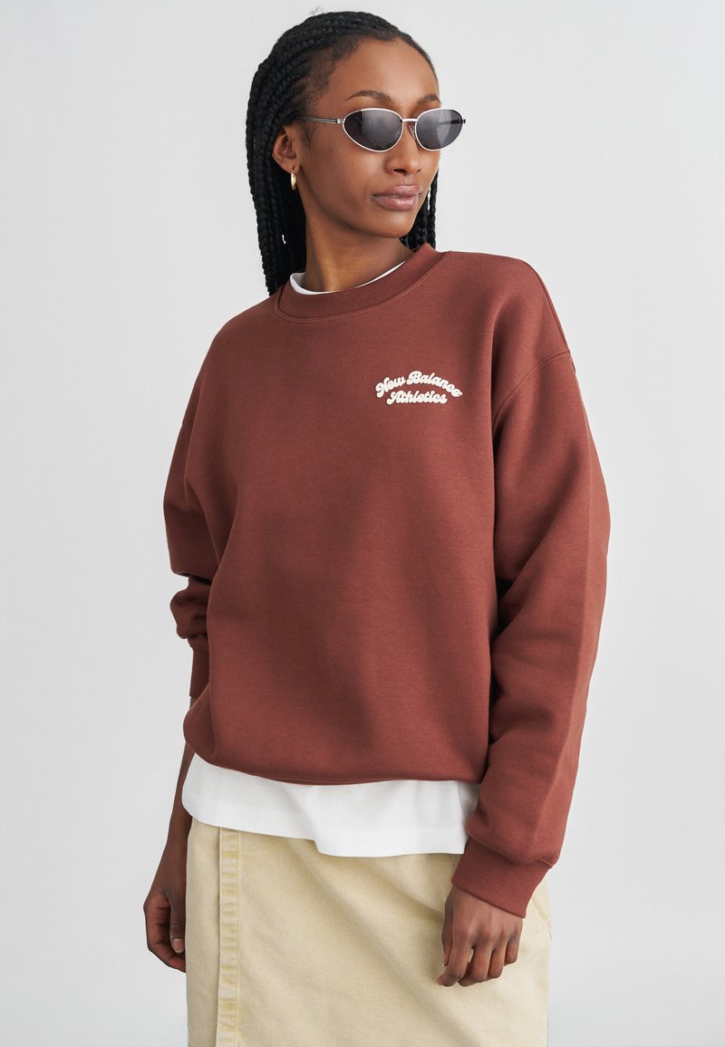 Sweatshirt castanho em algodão com o logótipo branco "New Balance Athletics", gola redonda com nervuras e corte solto, sobreposto a uma camisa branca e uma saia creme.