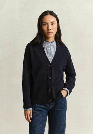 Woman with long dark hair wearing a navy buttoned cardigan over a ruffled collar shirt and blue jeans, standing with one hand in pocket.