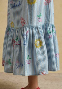 Blå- og hvidstribet nederdel med farverigt broderet sol, kirsebær, drikkevarer og ordet "le Soleil" ved bare ben og røde sko.