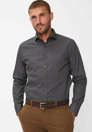 Chemise à motifs foncés avec un col pointu, présentant un design géométrique et des manches longues, associée à un pantalon chino marron et une ceinture marron.