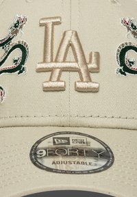 Căciulă de baseball bej cu logo-ul "LA" brodat în maro deschis. Prezintă modele de dragoni verzi și roșii. Eticheta ajustabilă New Era 9FORTY este vizibilă.