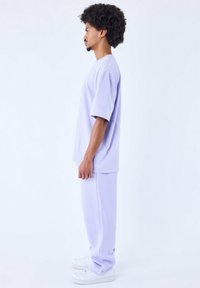 T-shirt oversize di un leggero viola e pantaloni jogger abbinati, realizzati in tessuto morbido. Il modello è in piedi di lato, indossando sneakers bianche.