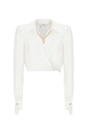 Blusa blanca, de manga larga y corta, hecha de una tela suave con un diseño envolvente. Presenta un collar de cadena dorada y acentos de botones en contraste.