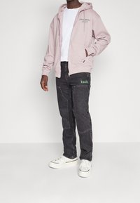 Sudadera con capucha y cremallera en rosa claro, con bolsillos frontales, camiseta blanca debajo, combinada con vaqueros negros de mezclilla con texto verde "ksubi" y elementos de patchwork.