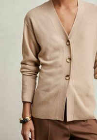 Beige kardigan vyrobený z měkké látky s véčkovým výstřihem, čtyřmi knoflíky, žebrovanými manžetami a dvěma předními kapsami. Nosený přes hnědé kalhoty.