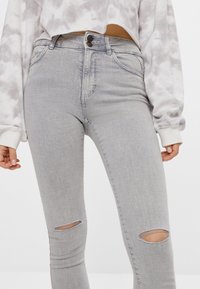 Jeans ajustados grises con dos desgastes en las rodillas, hechos de mezclilla, con una cintura de altura media y cierre de botón, combinados con una sudadera corta de tie-dye.