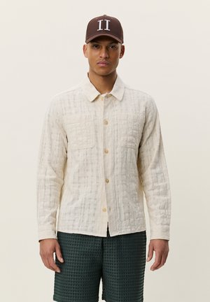 Homme portant une chemise à boutons texturée écrue, un short texturé foncé et une casquette marron avec deux lignes verticales blanches, debout devant un fond uni.
