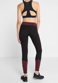 Kobieta ubrana w czarne legginsy w burgundowy wzór, czarny stanik sportowy i czarne sneakersy, stoi tyłem na jednolitym białym tle.