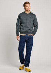 Sudadera gris oscura con cuello redondo, logo sutil en el pecho, combinada con una camisa de cuadros. Unos vaqueros azules y zapatillas amarillas completan el conjunto.