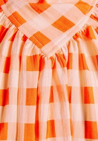 Tela cuadriculada en naranja y rosa claro con pliegues recogidos y un detalle triangular cosido en la parte superior.