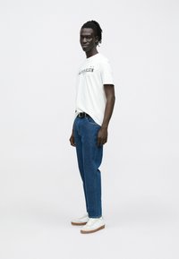 Mees dreadlockidega, kandmas valget Calvin Kleini T-särki, siniseid teksaseid, valgeid tossusid ja musta vööd, seistes ühevärvilise taustaga vastu.