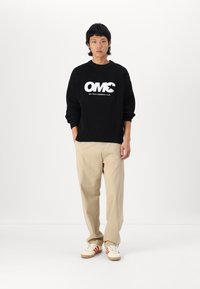 Suéter de punto negro con el logo blanco "OMC", pantalones rectos beige y zapatillas de crema con rayas rojas. Conjunto simple y casual.