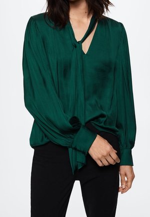 Mujer vistiendo una blusa de seda verde oscuro de manga larga con detalle de lazo en el cuello y pantalones negros, con la mano apoyada cerca de la cintura.