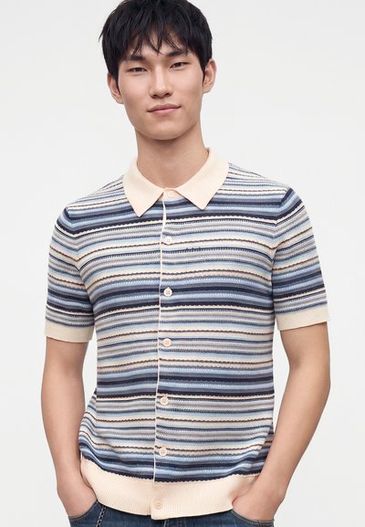 Jeune homme portant une chemise à manches courtes boutonnée avec des rayures horizontales bleues, blanches et beiges, debout les mains dans les poches devant un fond blanc.