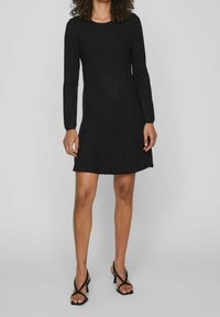 Donna che indossa un vestito a maniche lunghe a coste nere abbinato a sandali con tacco neri e fasce, in piedi contro uno sfondo grigio chiaro uniforme.