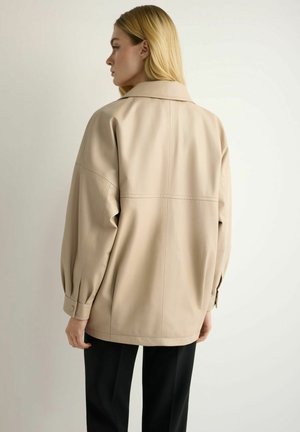 Kobieta o blond włosach, ubrana w beżową, oversize'ową kurtkę i czarne spodnie, widziana od tyłu na jednolitym jasnym tle.